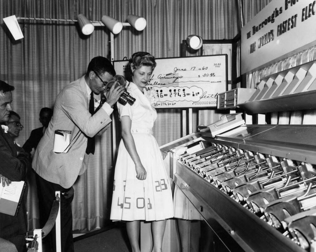 Image description: Vintage image of two people standing before a printing press; one points a film camera at the machine. End of alt text.
