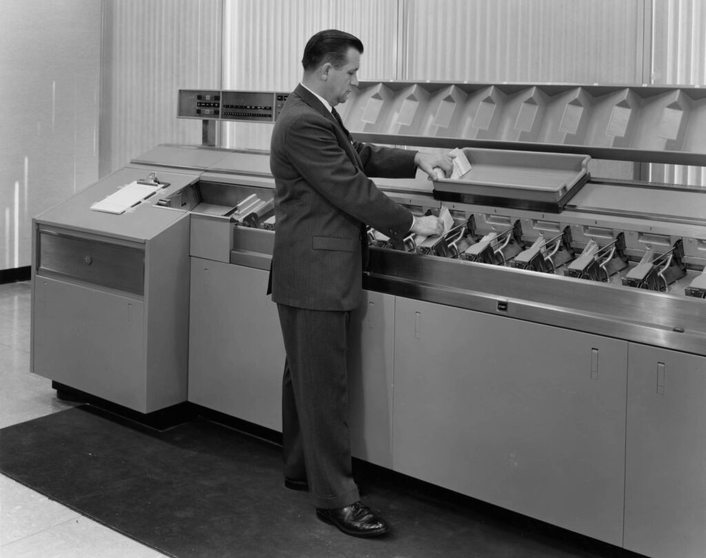 Image description: Vintage image of a person in a suit collecting checks from a printing machine. End of alt text.