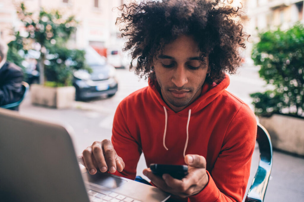 Image description: Closeup of a person in a red hooded sweatshirt sitting by an open laptop looking at the phone in their hand. End of alt text.